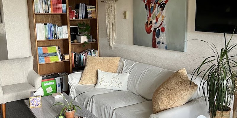 A cozy living space with a soft-cushioned couch, colorful artwork, bookshelves, indoor plants, and a coffee table, creating a welcoming vibe.