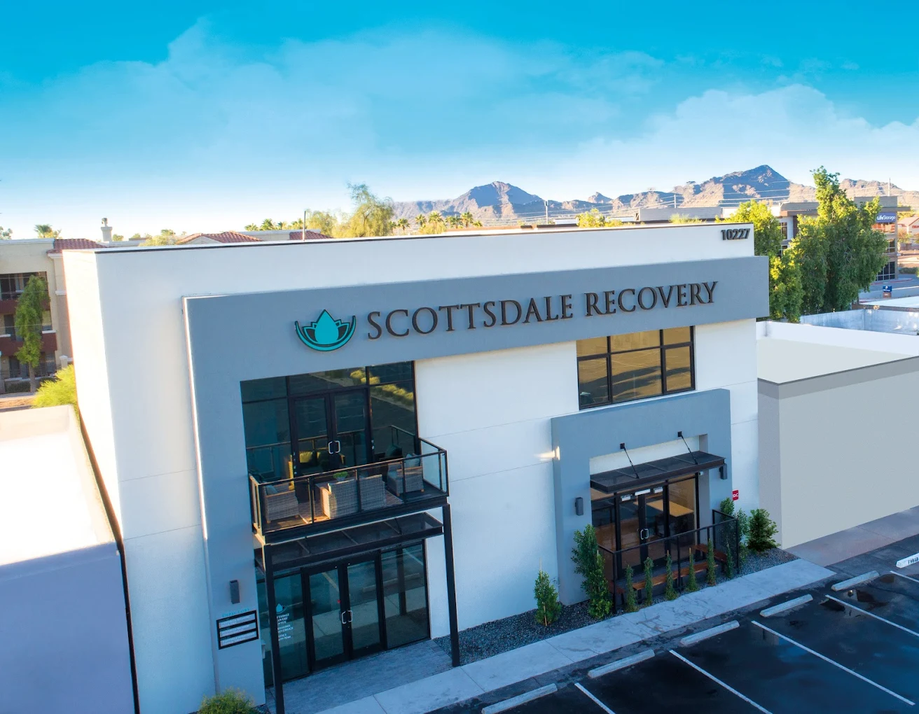 The front exterior of Scottsdale Recovery, a modern two-story building with large glass doors, balconies, and landscaped entryways against a backdrop of distant mountains