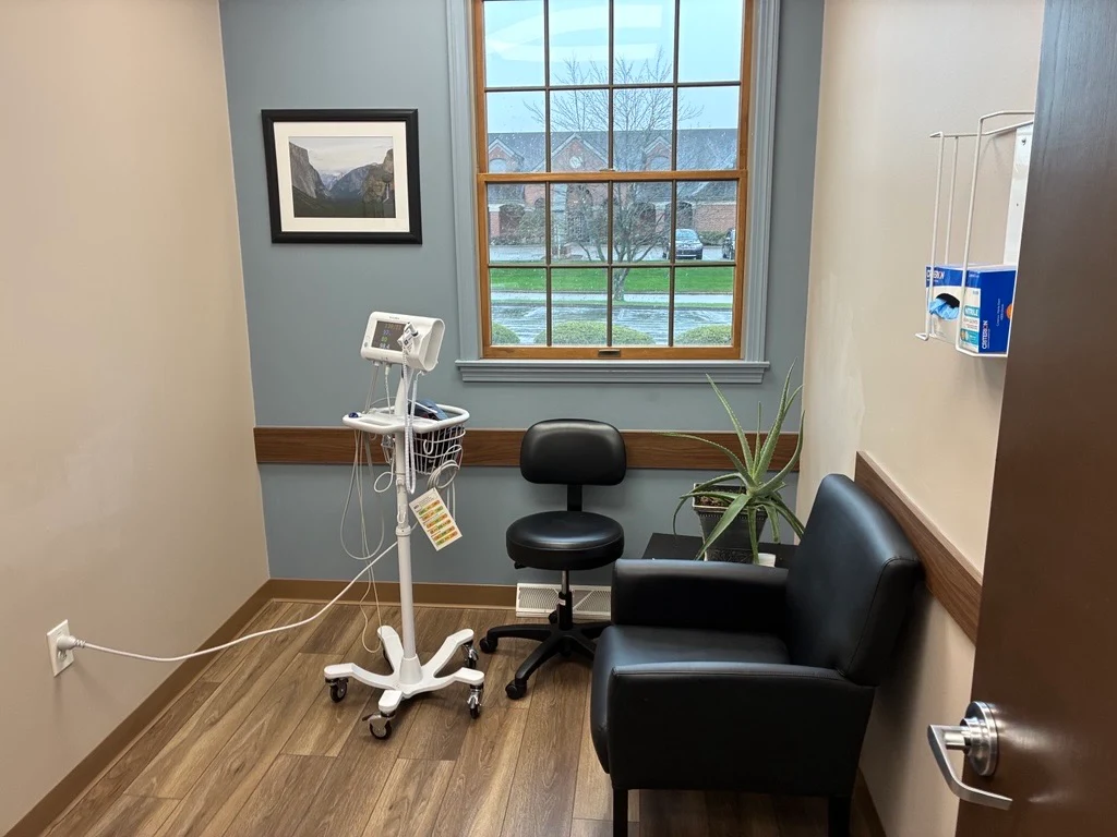 Clinical room with vital-sign monitoring equipment, patient seating, large window, wall-mounted glove dispenser, and wood-look flooring