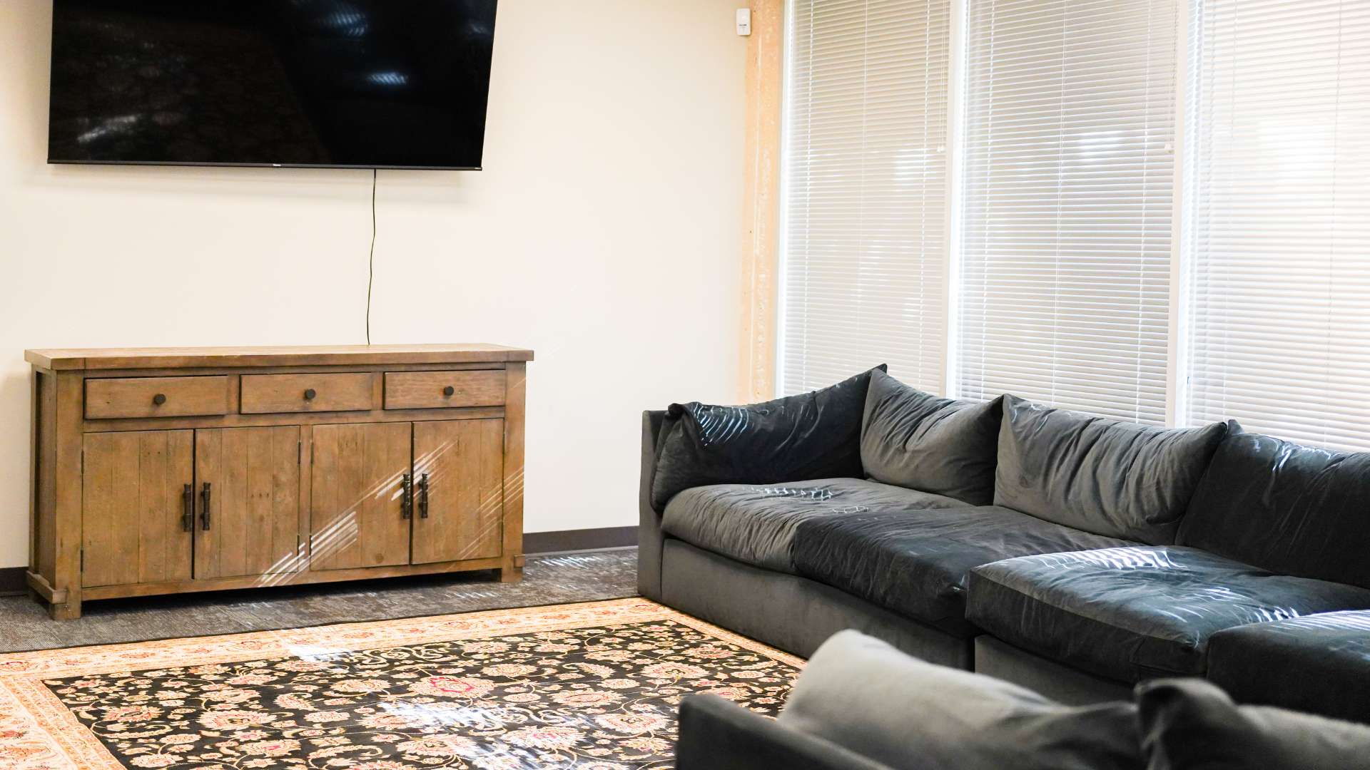 Comfortable TV lounge with a large dark sectional sofa, wall-mounted television, wooden console, patterned area rug, and bright windows
