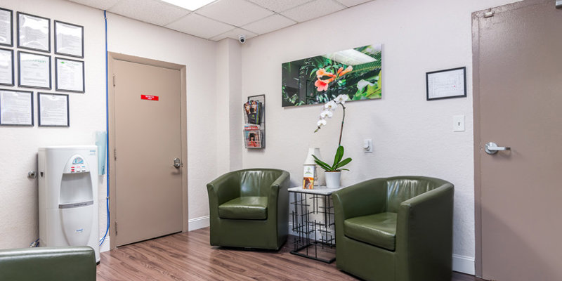 A cozy waiting area with comfortable green chairs, a small table holding a potted orchid, framed artwork, and a water dispenser for added convenience.