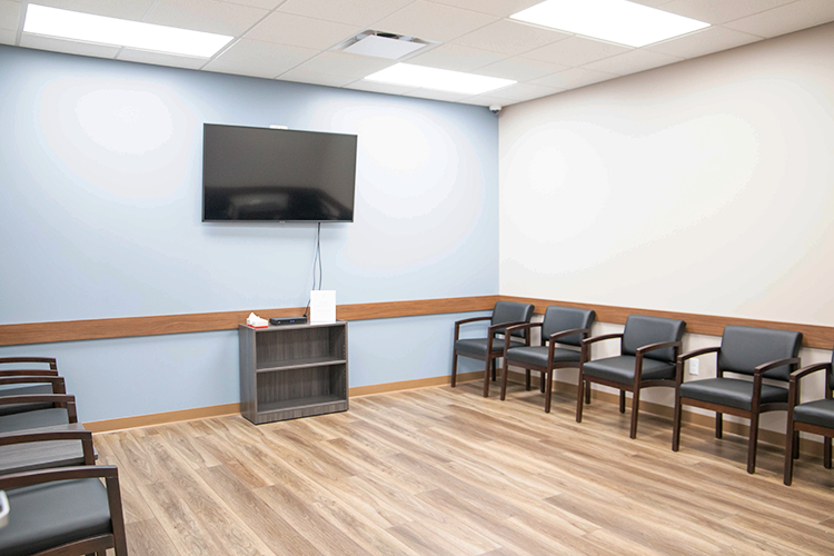 Treatment and waiting room with black chairs arranged along the walls, wall-mounted television, and wood-look flooring