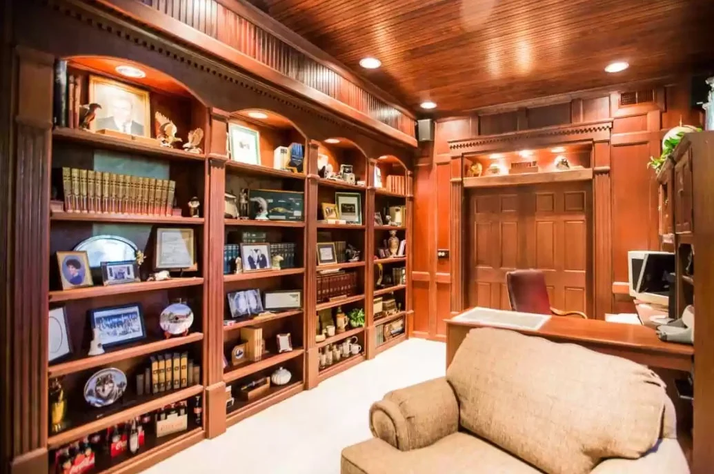 Wood-paneled library and office with built-in bookshelves, desk, computer, armchair, framed photographs, and recessed lighting