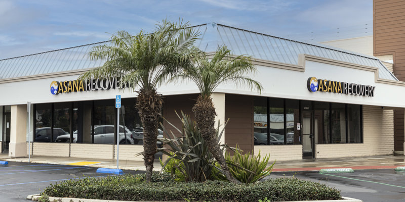 A clean, modern building with large windows and palm trees outside, featuring the Asana Recovery logo on the storefront.