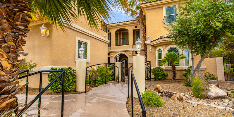 A welcoming Mediterranean-style home with a landscaped entrance, vibrant greenery, palm trees, and a smooth pathway leading to the front door.