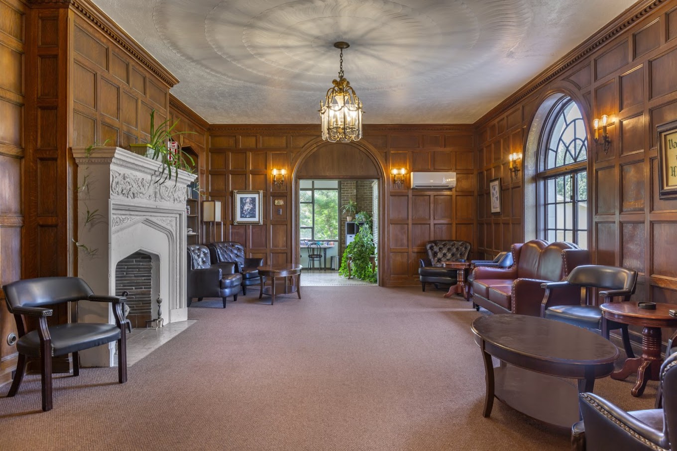 A classic-style living room with wooden panel walls, a fireplace, leather seating, and elegant chandeliers.