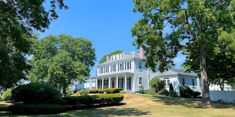 A grand white building surrounded by lush greenery and tall trees on a sunny day, creating a peaceful and inviting atmosphere.