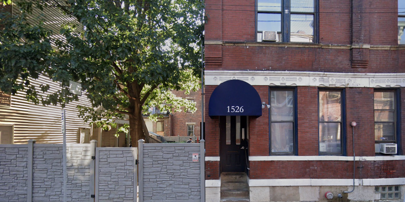 A welcoming brick building with large windows and a tree-lined street, featuring an arched blue awning and the address number 1526.