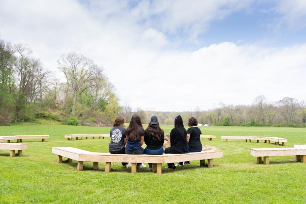 Group of women seated on wooden benches arranged in a circular formation in an open grassy field