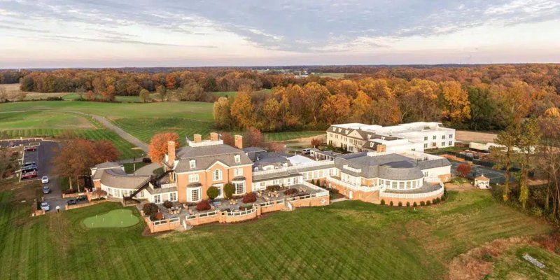 Aerial view of an expansive estate surrounded by autumn foliage, featuring a grand building, landscaped lawns, and scenic countryside.