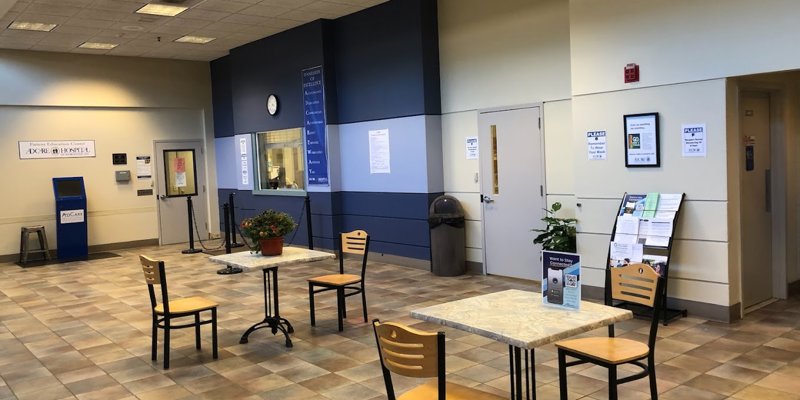 A spacious lobby with tiled floors, light wooden chairs around marble-top tables, and vibrant potted plants.