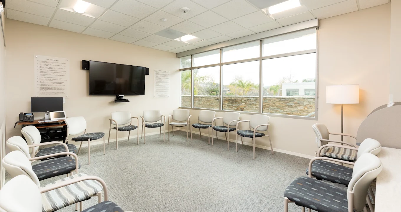 Therapy room with chairs arranged in a circle, a wall-mounted television, and large windows providing natural light