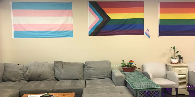  A cozy room with a gray sectional sofa, LGBTQ+ and transgender pride flags displayed on the wall, fostering an inclusive, welcoming atmosphere.