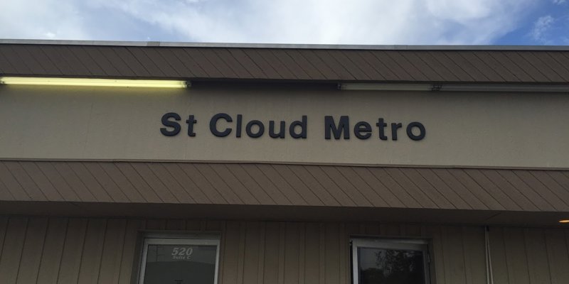 Close-up of the "St Cloud Metro" sign on a beige building with two glass doors below, under a partly cloudy sky, offering a simple exterior view.