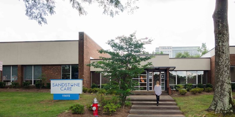 A person walks toward the modern, welcoming entrance of Sandstone Care, surrounded by greenery and a well-maintained landscape.