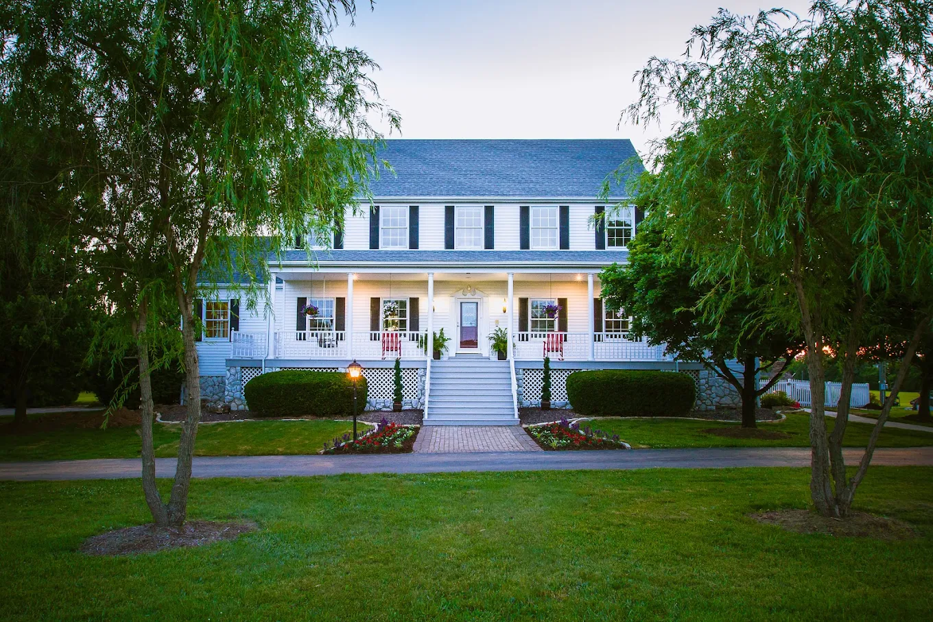 Colonial-style white house with a front porch, black shutters, landscaped garden, and evening lighting