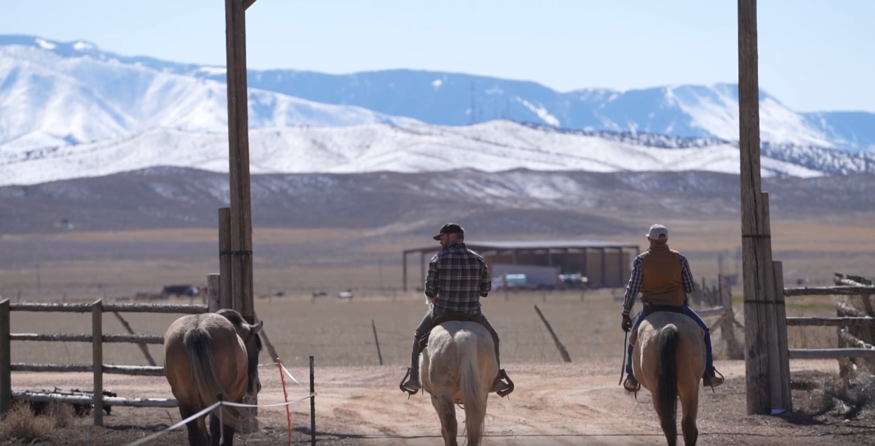 Two men on horseback ride away under a large wooden ranch archway.