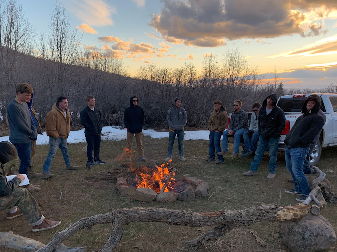 Group of men gathered around a campfire at dusk near snowy trees.