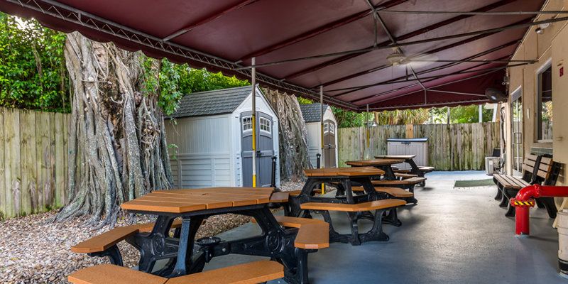 A shaded outdoor seating area with several picnic tables and benches, surrounded by a wooden fence and greenery, creating a peaceful atmosphere.