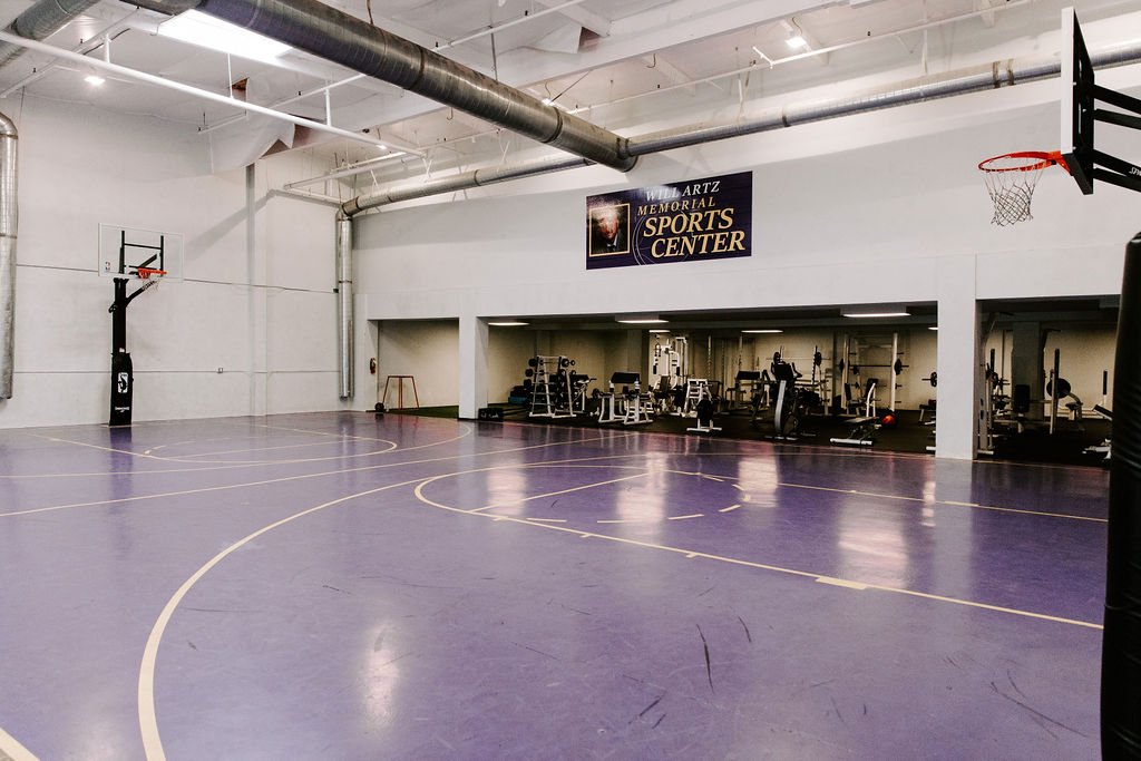 Indoor basketball court with purple flooring, weight training area, and "Will Artz Memorial Sports Center" banner