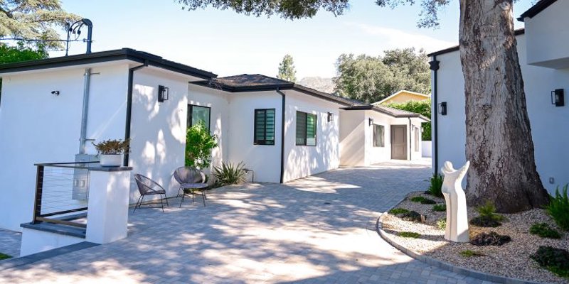 A tranquil, tree-shaded courtyard features modern white buildings, cozy outdoor seating, and thoughtful landscaping, offering a peaceful retreat.