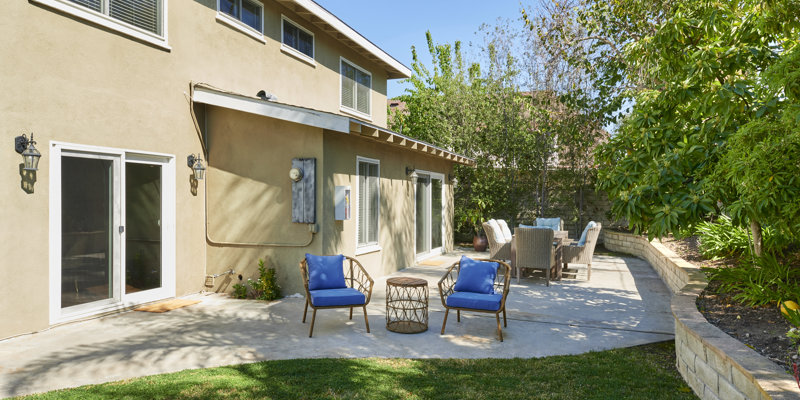 A sunny backyard patio with blue cushioned chairs and a cozy outdoor dining area, surrounded by lush greenery and trees, perfect for relaxation.