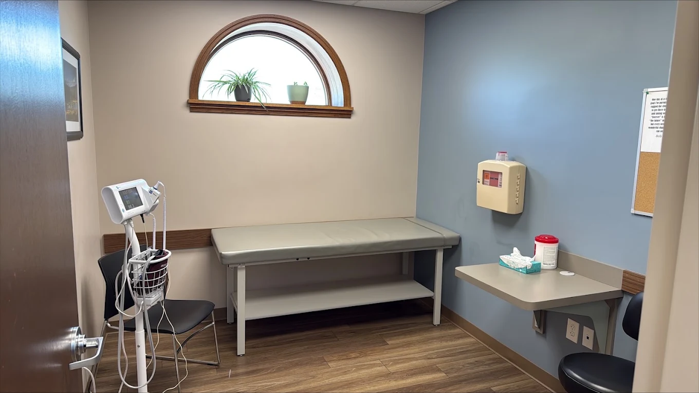 Clinical exam room with examination table, vital-sign monitor, medical supplies, chairs, and an arched window