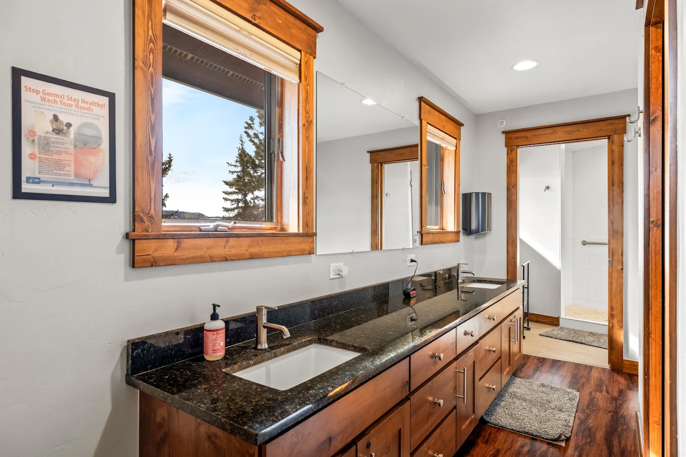 Bright bathroom with a long granite vanity, double sinks, mirrors, wood cabinetry, and walk-in shower