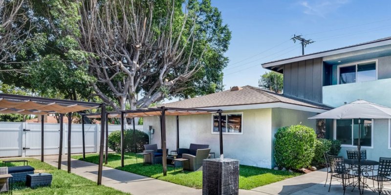 A cozy outdoor seating area with shaded pergolas, comfortable chairs, and umbrella-covered tables, surrounded by lush greenery and trees.