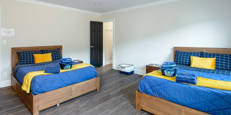 A bright, spacious bedroom with two neatly made twin beds, featuring blue and yellow bedding, wood furniture, and ample natural light.