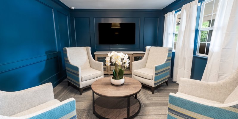 A cozy seating area with elegant armchairs and a wooden table, enhanced by bright windows and a soothing blue accent wall with a TV.