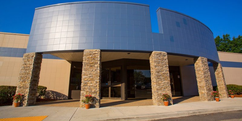 A modern building with a sleek curved facade featuring gray tiles and stone columns at the entrance. Flowerpots line the walkway for a welcoming touch.