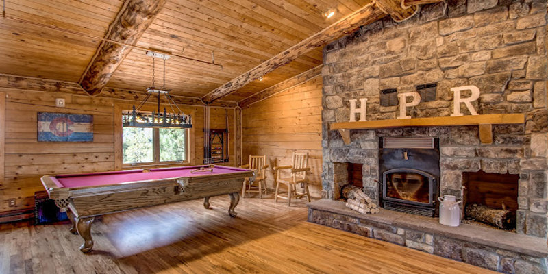 A rustic game room with a wooden pool table, stone fireplace, and exposed log beams, offering a cozy and entertaining space for relaxation.