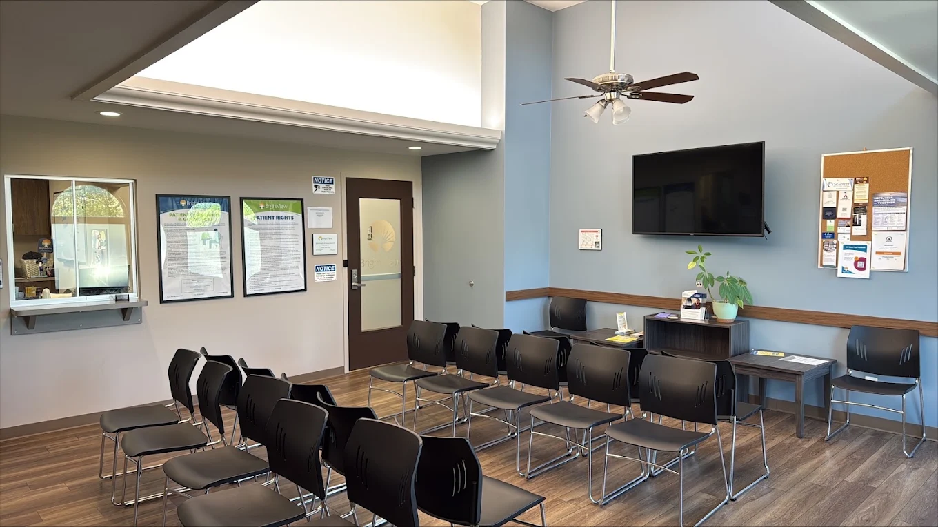 BrightView Health lobby with rows of black chairs, reception window, wall-mounted television, bulletin board, and high ceiling
