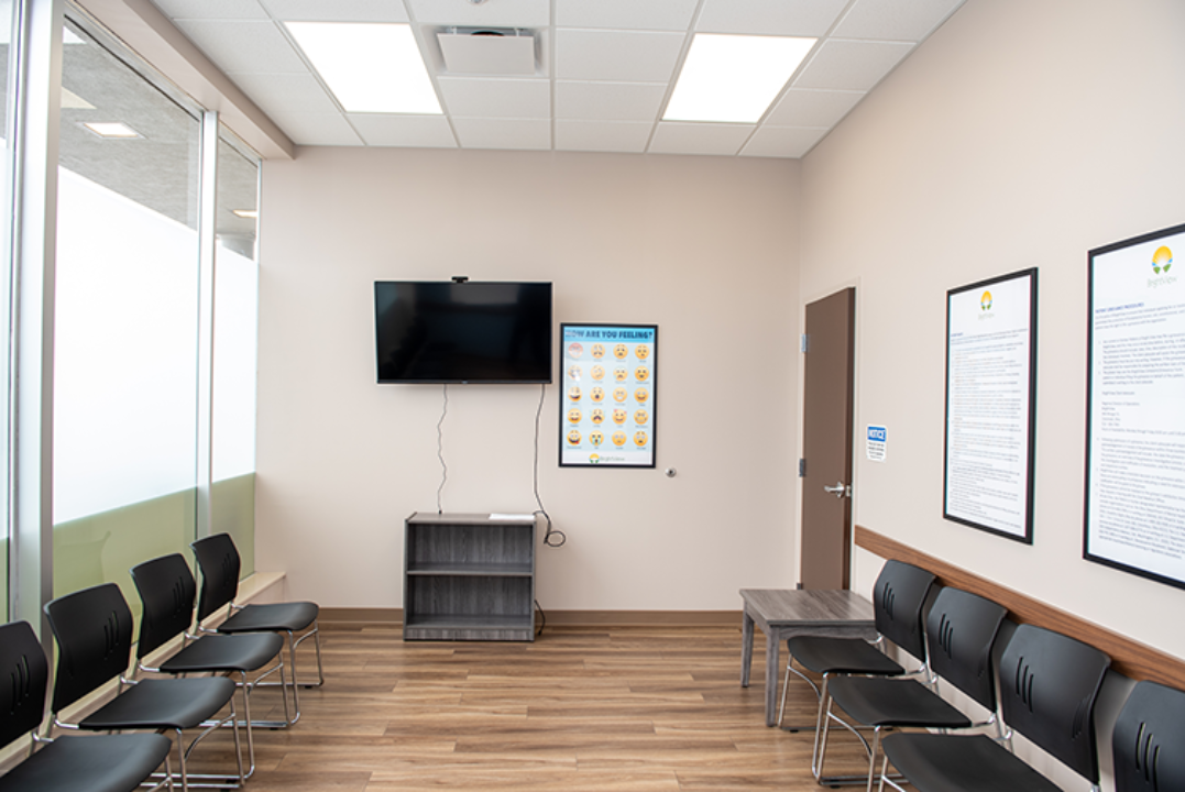 BrightView Health waiting area with rows of black chairs, wall-mounted television, informational posters, large windows, and wood-look flooring