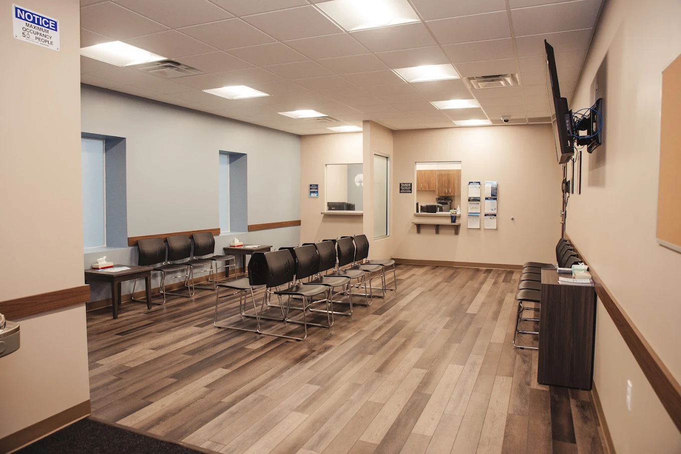 Spacious BrightView Health waiting area with rows of black chairs, service windows, wall-mounted television, wood-look flooring, and neutral walls with blue accents