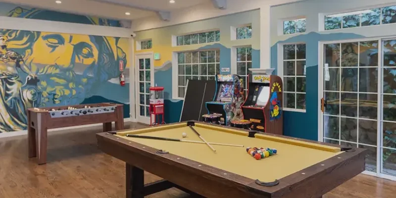 A vibrant game room featuring a pool table, foosball, arcade games, and colorful wall art. Large windows offer plenty of natural light.