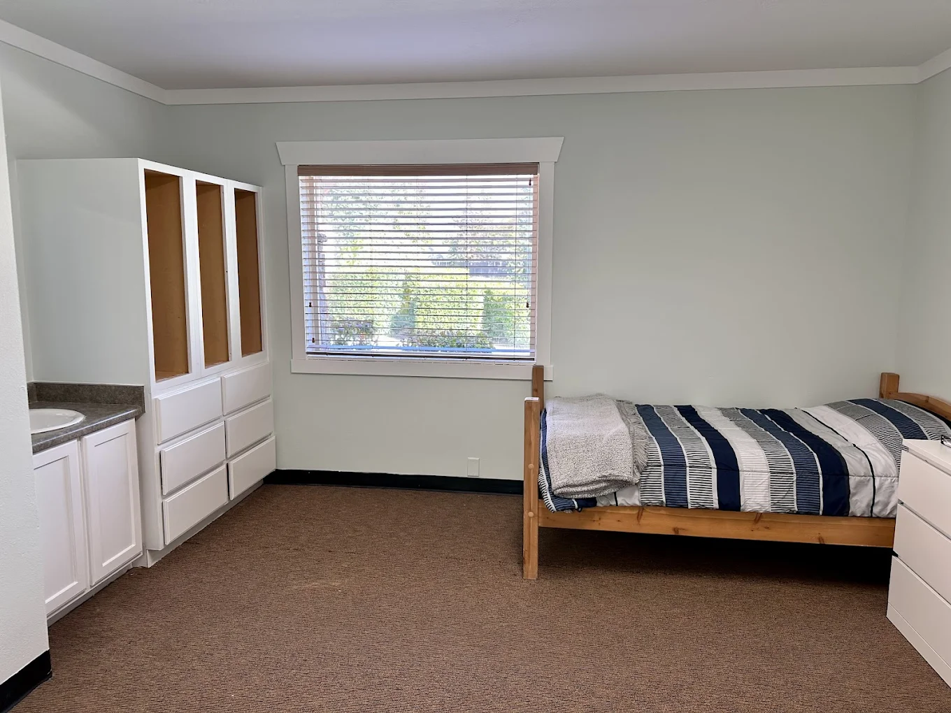 Simple bedroom with single bed, built-in storage, sink, and window view at New Roads Behavioral Health