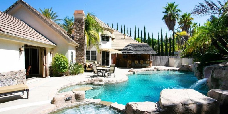 A stunning backyard oasis featuring a sparkling pool, waterfall, jacuzzi, tropical landscaping, and a cozy outdoor seating area for relaxation.