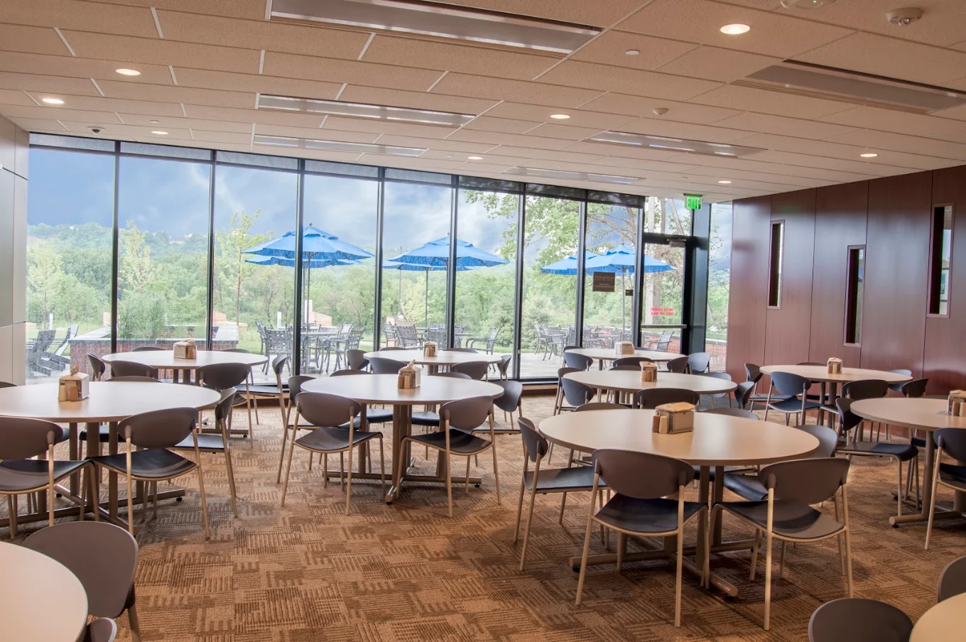 Spacious dining area with round tables and chairs overlooking an outdoor patio and wooded landscape