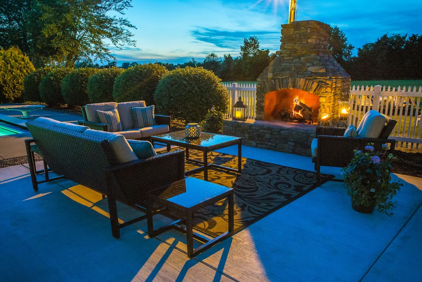 Outdoor seating area with cushioned furniture and a stone fireplace lit at dusk