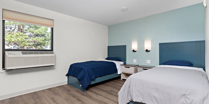 A cozy bedroom with two single beds, soft blue headboards, and wooden flooring. The large window allows natural light to create a calming atmosphere.