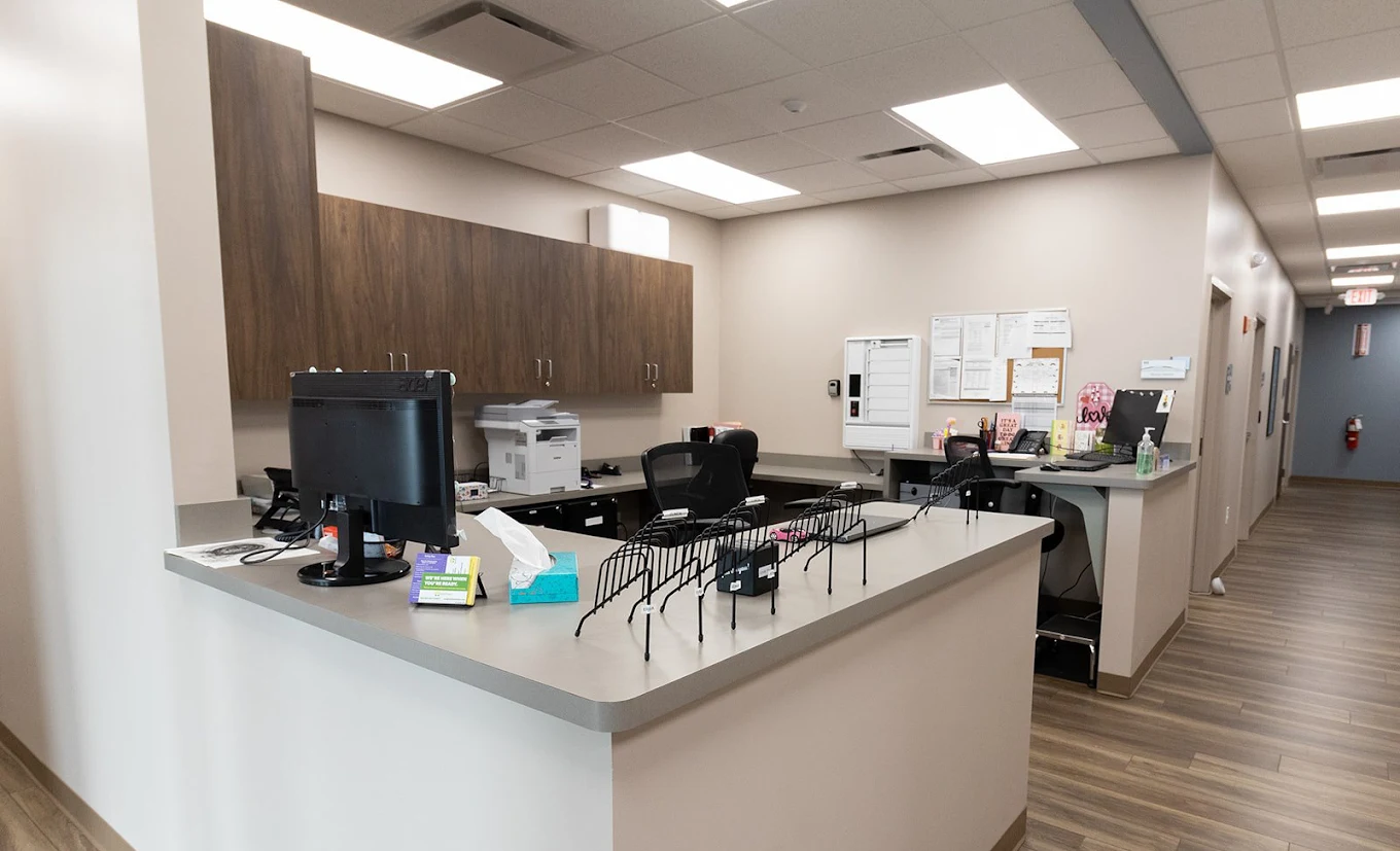 BrightView Health staff workstation with wraparound counter, computers, office chairs, printer, overhead cabinets, document organizers, and adjacent hallway