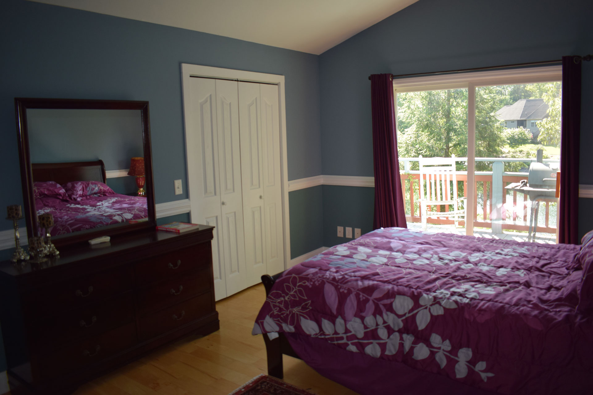 Bedroom with blue walls, purple floral bedding, wood dresser, large mirror, hardwood flooring, and sliding glass doors opening to a deck