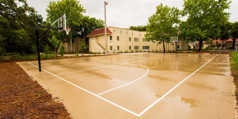 A freshly paved outdoor basketball court with clear white lines, surrounded by lush greenery and a peaceful building in the background.