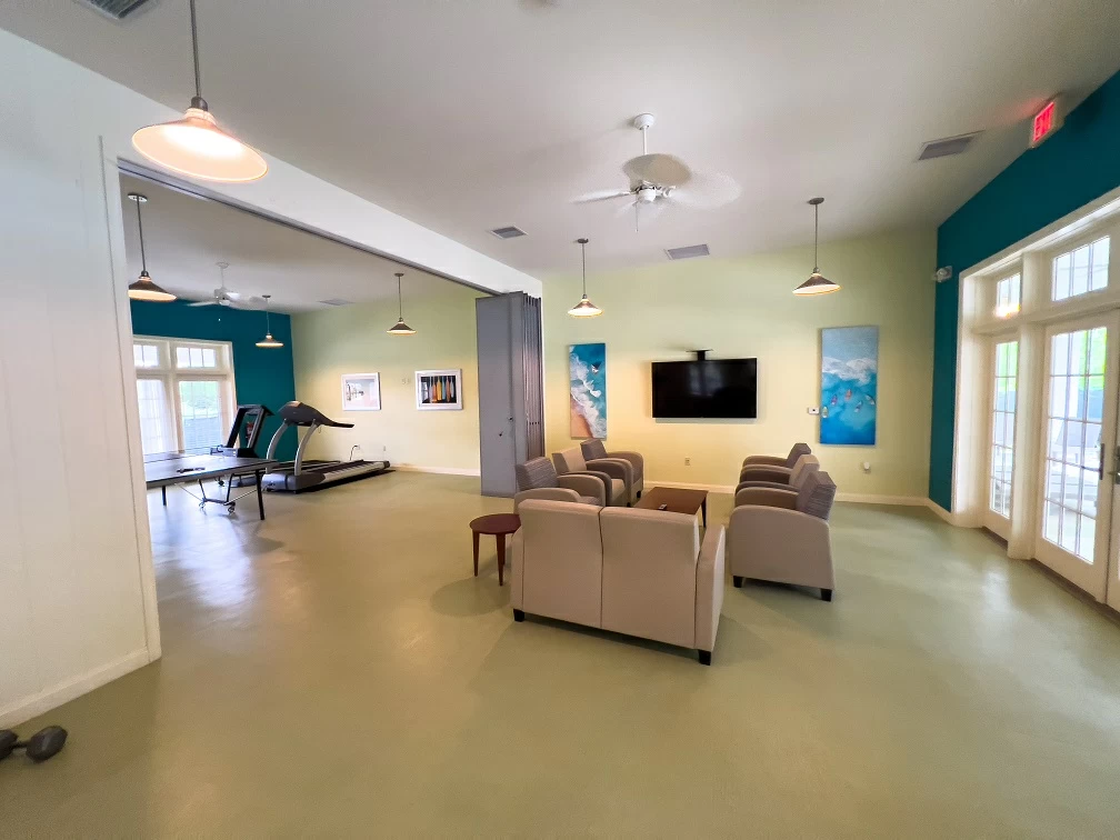 Open wellness lounge with seating area, treadmill, television, and large windows inside recovery center