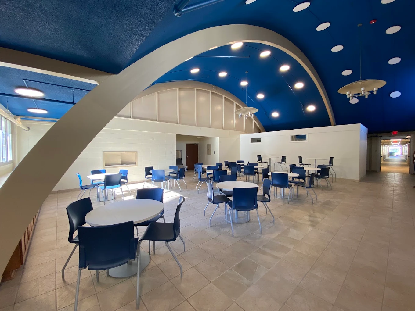 Spacious recreational and dining area with round tables, blue chairs, high arched ceilings, and overhead lighting