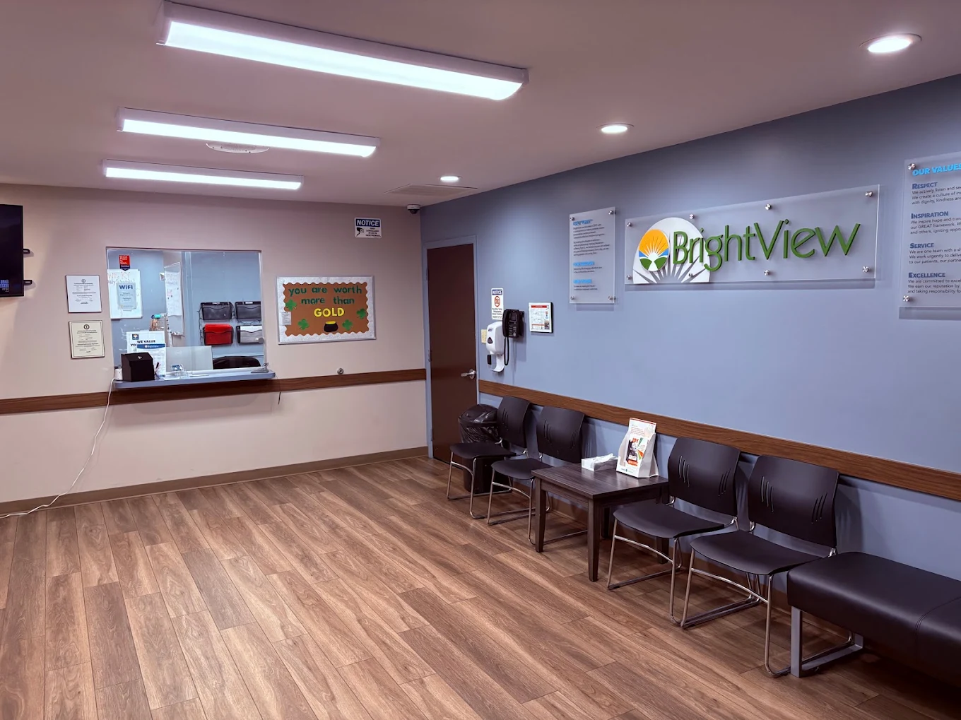 BrightView Health reception and waiting area with check-in window, black chairs, BrightView wall logo, informational displays, and wood-look flooring