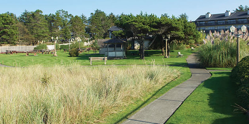 A peaceful garden with a winding path, gazebo, and shaded benches surrounded by tall grasses and trees, leading to a charming resort.