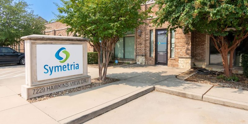 A welcoming facility entrance with a prominent "Symetria" sign, surrounded by greenery and a well-maintained exterior, creating an inviting atmosphere.